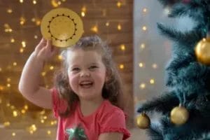 Meisje houdt lachend Penta Power Gold Tag naast kerstboom — cadeau voor een gezonde, ondersteunende thuisomgeving